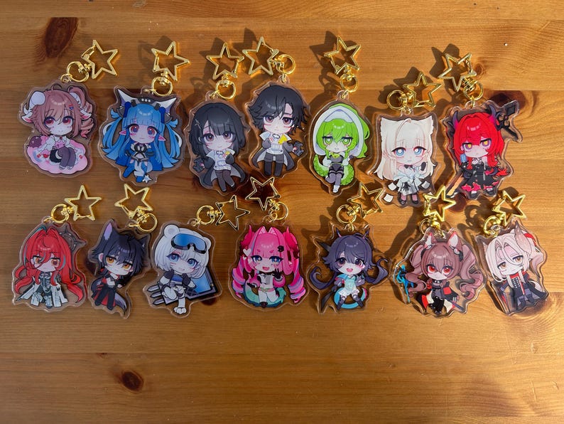 May include: Collection of anime-style character keychains with gold star-shaped clasps. Each keychain features a unique character design with vibrant colors and detailed artwork. The keychains are arranged on a wooden surface.
