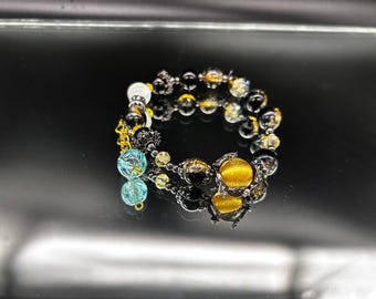 May include: A beaded bracelet featuring a mix of black, gold, and clear beads, including a prominent yellow cat's eye bead and two dangling light blue crackle glass beads. The bracelet is accented with small gold-toned metal spacers and charms.