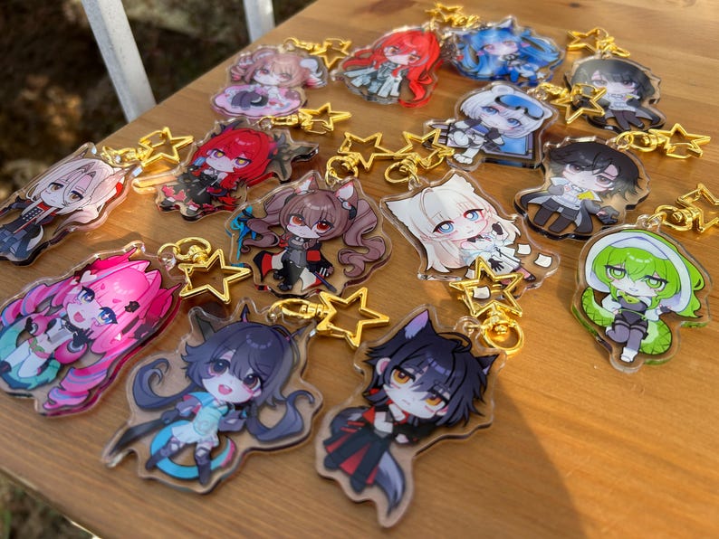 May include: Collection of anime-style character keychains with gold star-shaped clasps. The keychains feature various characters with colorful hair and outfits. The keychains are displayed on a wooden surface.