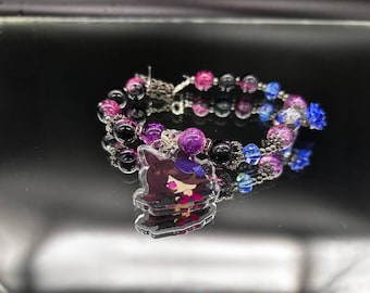 May include: A beaded bracelet featuring a mix of black, purple, and blue beads with silver-toned spacers. A clear acrylic charm depicting a stylized character with dark hair and a purple hat hangs from the bracelet, which includes a metal clasp.