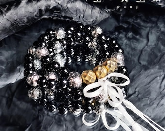 May include: A multi-strand beaded bracelet featuring glossy black spherical beads, textured silver-toned metallic beads, and three matte gold-toned hammered beads. A white ribbon is tied into a bow on the side. The text 望 - 長 生 劫 w a n g is visible.