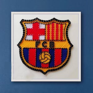 Puede incluir: Una pieza de arte de hilo enmarcada que presenta el escudo del FC Barcelona. El escudo, con forma de escudo, utiliza hilos rojos, azules, amarillos y blancos. El diseño incluye una cruz, rayas y un balón de fútbol. El marco es blanco, sobre un fondo azul.