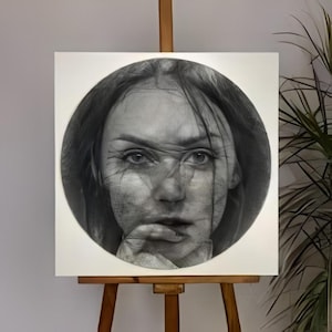 Pode incluir: Uma pintura em preto e branco de um retrato do rosto de uma mulher, emoldurada em um círculo. A obra de arte é apresentada em uma tela branca, apoiada por um cavalete de madeira. A expressão da mulher é intensa, com rugas visíveis e uma pose pensativa.