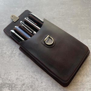 May include: A brown leather pen case, open to show several pens. The case is rectangular with a flap closure secured by a small metal clasp. A metal ring is attached to the front. The pens are silver and black.