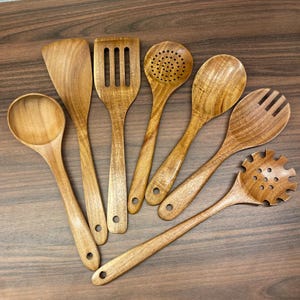 Può includere: Una collezione di sette utensili da cucina in legno, tra cui una spatola, un cucchiaio scanalato e cucchiai da portata. Gli utensili sono realizzati in legno marrone chiaro con venature visibili e fori nei manici.
