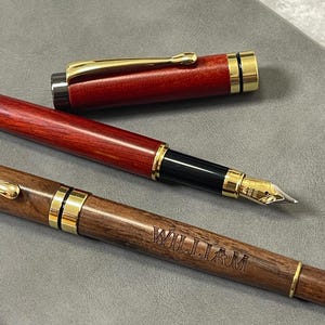 Puede incluir: Dos plumas estilográficas con cuerpo de madera y detalles dorados. Una es de color rojo oscuro, la otra marrón medio. La pluma marrón tiene grabado el nombre "WILLIAM". Las plumas se muestran sobre una superficie gris.