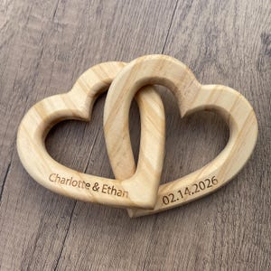 May include: Two interlocking light-colored wooden heart shapes with a natural grain pattern. The left heart is engraved with the text Charlotte & Ethan, and the right heart is engraved with the date 02.14.2026 in a dark, clear font.