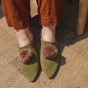 Puede incluir: Mules de ante verde oliva con punta y un gran pompón redondo de color malva. Los zapatos se llevan con pantalones color óxido. Los zapatos están sobre una alfombra estampada.