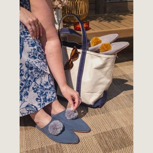 Puede incluir: Un par de zapatillas de ante azul con pompones grises, junto a un bolso tote de lona con ribete azul marino. El bolso contiene zapatillas grises con pompones amarillos y gafas de sol. La escena se desarrolla sobre una estera tejida.