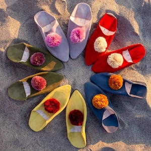 Puede incluir: Una colección de coloridas zapatillas de ante dispuestas en círculo sobre arena. Las zapatillas vienen en varios colores, incluyendo rojo, azul, amarillo, verde y lavanda, cada una adornada con un pompón a juego. Las zapatillas tienen un diseño de punta.