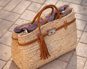 May include: A woven straw tote bag with brown leather accents. The bag features braided handles, a leather strap with a metal clasp, and a decorative tassel. The interior lining is a light purple color.