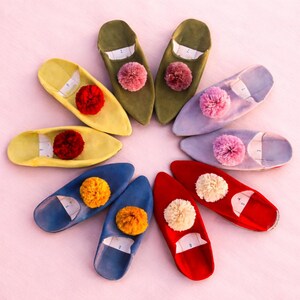 Puede incluir: Una colección de coloridas y suaves zapatillas dispuestas en un patrón circular. Las zapatillas vienen en varios colores, incluyendo amarillo, verde, azul, rojo y morado, cada una adornada con un pompón esponjoso. Las zapatillas parecen estar hechas de tela suave.