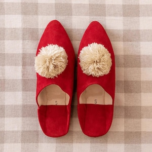 Puede incluir: Un par de zapatillas de ante rojas con un pompón color crema en cada punta. Las zapatillas tienen un diseño de talón abierto y son de talla 5. Las zapatillas se muestran sobre un fondo de tela a cuadros.