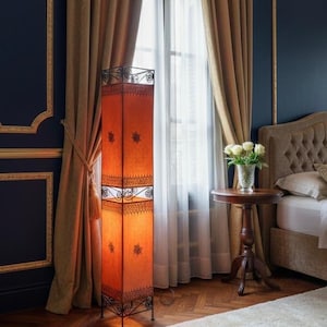 May include: An orange rectangular floor lamp with a decorative metal frame. The lamp has a patterned design and stands tall in a room with dark blue walls, gold trim, and tan curtains. A small round table and bed are also visible.
