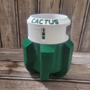 May include: A green and white cactus-shaped planter with the word "CACTUS" printed on the top. The planter has a textured surface and a unique design, perfect for succulents or small plants. The planter is made of a durable material.