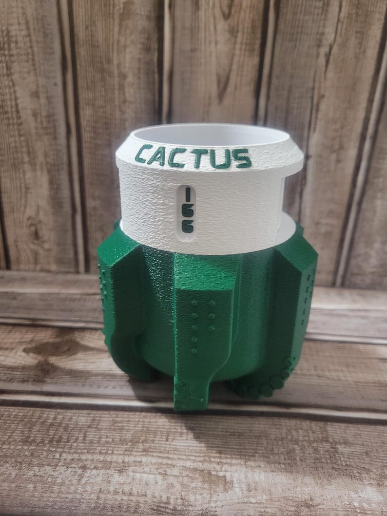 May include: A green and white cactus-shaped planter with the word "CACTUS" printed on the top. The planter has a textured surface and a unique design, perfect for succulents or small plants. The planter is made of a durable material.