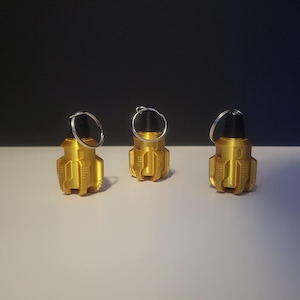May include: Three gold-colored, miniature grenade keychains with black tops and silver key rings. The keychains are arranged in a row on a white surface, with a dark background.