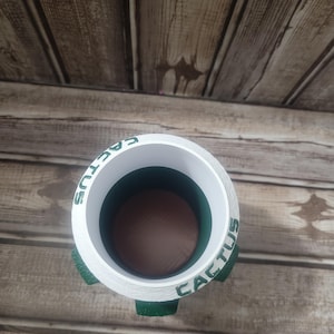 May include: A white cylindrical planter with the word "CACTUS" in green lettering around the rim. The planter has three green, rounded feet. The interior is dark brown and green. The planter is sitting on a wooden surface.