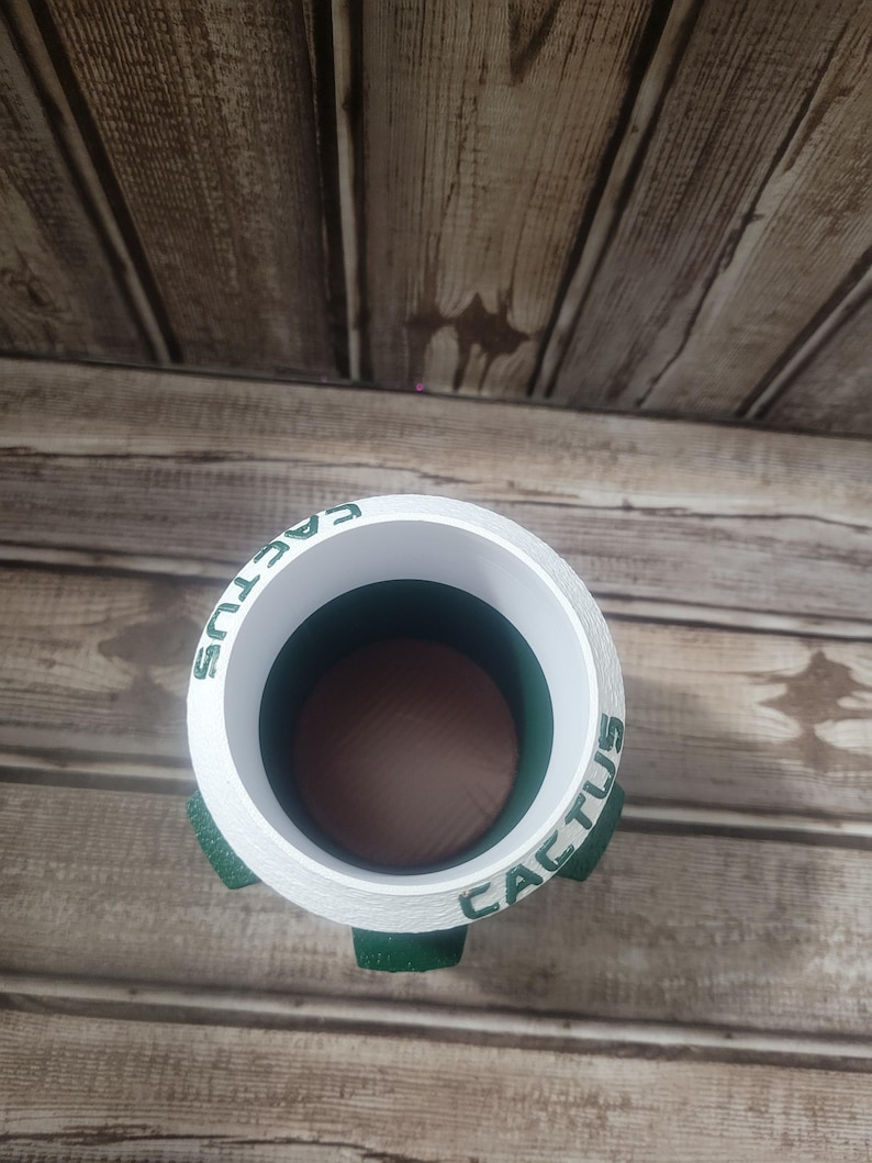 May include: A white cylindrical planter with the word "CACTUS" in green lettering around the rim. The planter has three green, rounded feet. The interior is dark brown and green. The planter is sitting on a wooden surface.
