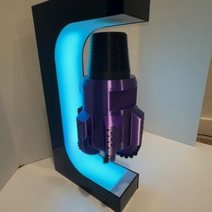 May include: A purple drill bit model suspended within a black C-shaped stand that features an integrated blue LED light strip along its inner curve, illuminating the metallic purple object from behind while it floats in the center of the frame.