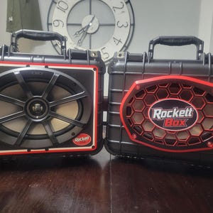 May include: Two black portable speaker cases with carrying handles. The left case features an oval speaker with a black grille and a small Rockett Box logo. The right case has a hexagonal red and black grille with the text Rockett Box in the center.