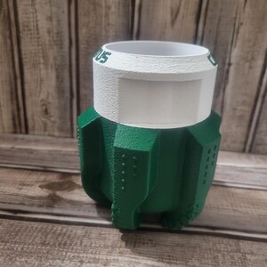 May include: A green and white 3D-printed planter. The planter has a textured green base with a white rim and the text "US" visible. The planter is designed with a unique, geometric shape.