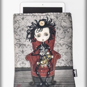 Puede incluir: Una funda gris para tableta con un personaje parecido a una muñeca con cabello negro y un atuendo negro, sentado en una silla roja. El personaje sostiene un pequeño osito de peluche. El fondo tiene un patrón de damasco gris.