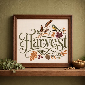 May include: A framed cross-stitch embroidery featuring the word Harvest in dark green cursive lettering, surrounded by autumn-themed motifs including wheat stalks, a small bird, a maple leaf, an oak leaf, berries, and an acorn on fabric.