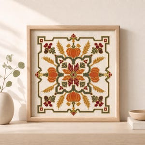 Könnte beinhalten: Eine quadratische Kreuzstichstickerei in einem hellen Holzrahmen mit einem symmetrischen herbstlichen Muster. Das Design zeigt orangefarbene Kürbisse, goldene Weizenähren, dunkelgrüne Blätter und kleine rote Beeren in einem geometrischen Raster.