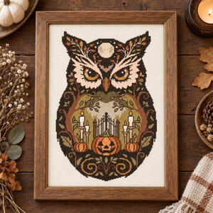 May include: A framed cross-stitch embroidery featuring a stylised owl with dark brown and black plumage. The owl's chest contains a scene with a jack-o'-lantern, a wrought iron gate, and lit candles, all set against a cream-coloured fabric background.