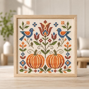 May include: A square framed cross-stitch embroidery featuring two orange pumpkins at the bottom, flanked by symmetrical floral motifs and two blue birds with orange wings facing inward toward a central red and blue stylized flower design.