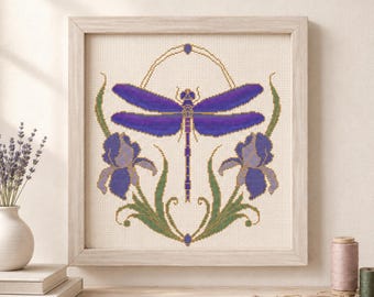 May include: A framed cross-stitch embroidery featuring a central purple dragonfly with extended wings, flanked by two purple iris flowers with green stems and leaves, all set against a cream-coloured fabric background within a light wood frame.