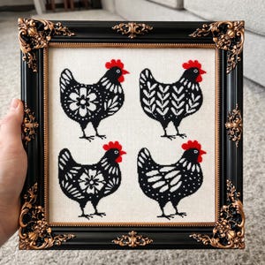 Puede incluir: Arte de punto de cruz enmarcado con cuatro gallinas en blanco y negro con crestas rojas. Las gallinas tienen varios patrones decorativos. La obra de arte está en un marco negro con detalles dorados. Una persona sostiene el marco.