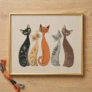 May include: A framed cross-stitch artwork depicting five stylized cats. The cats are in black, tan, orange, grey, and brown. Each cat has a unique design, with star and leaf patterns. The frame is gold-coloured.