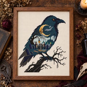 May include: A framed cross-stitch embroidery featuring a dark blue raven perched on a branch. The bird's body contains a scene of a cemetery with a glowing lantern, a gate, and a yellow crescent moon set against a dark blue night sky.