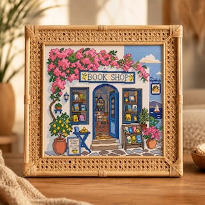 Op de afbeelding: Een kruissteekborduurwerk van een boekwinkel in mediterrane stijl in een licht houten rotan lijst. Het ontwerp toont roze bougainville, een citroenboom en borden met de tekst BOOK SHOP en SO MANY BOOKS SO LITTLE TIME met blauwe deuren.