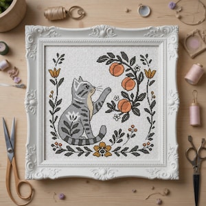May include: Cross-stitch artwork featuring a grey tabby cat and peach branches, framed in white. The design includes a floral border with orange and yellow details. The frame is ornate.