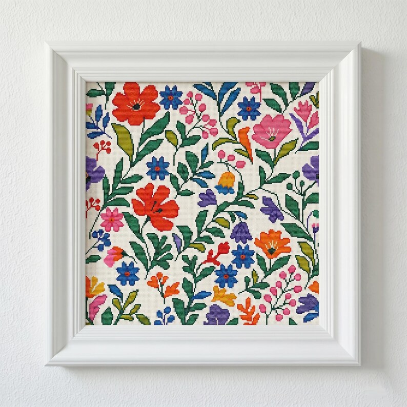 May include: A framed floral cross-stitch pattern. The design features a variety of colorful flowers, including red, pink, blue, and orange, set against a cream background. The flowers are surrounded by green leaves and stems. The frame is white.