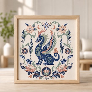 May include: A square framed cross-stitch embroidery featuring a central dark blue dragon with wings, surrounded by a symmetrical floral wreath in shades of peach, sage green, and navy blue, set against a cream-colored fabric background.