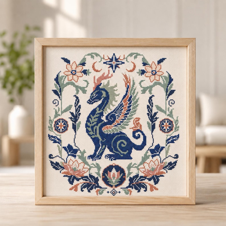 May include: A square framed cross-stitch embroidery featuring a central dark blue dragon with wings, surrounded by a symmetrical floral wreath in shades of peach, sage green, and navy blue, set against a cream-coloured fabric background.