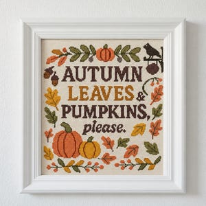 以下が含まれることがあります： 「AUTUMN LEAVES & PUMPKINS, please.」の文字が入った、額装されたクロスステッチアート。カボチャ、葉、ドングリが秋の色で描かれています。黒い鳥が枝に止まっています。アートワークは白いフレームに入っています。