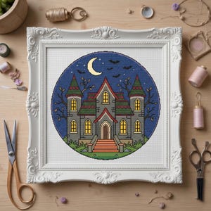 May include: A framed cross-stitch design featuring a spooky haunted house scene. The house is gray with green and red accents, set against a dark blue night sky with a crescent moon and bats. The frame is white.