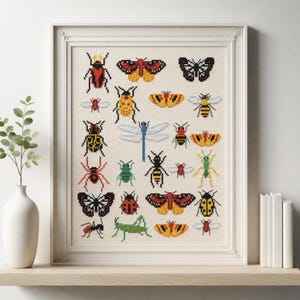 Könnte beinhalten: Ein gerahmtes Kreuzstichbild mit einer Sammlung farbenfroher Insekten, darunter Schmetterlinge, Bienen und Käfer. Das Kunstwerk wird in einem weißen Rahmen präsentiert und steht auf einem Holzregal. Die Insekten sind in einem pixeligen Stil dargestellt.