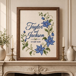 May include: A framed cross-stitch embroidery featuring the text Taylor and Jackson and 15 SEP 2026 in dark blue script. A floral wreath of blue clematis flowers and green leaves curves along the right side of the cream-colored fabric.