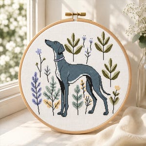 May include: A cross-stitch embroidery hoop featuring a greyhound dog design. The dog is surrounded by stylized plants and flowers in shades of green, blue, and yellow. The embroidery is set in a wooden hoop.
