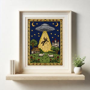 May include: Framed cross-stitch art depicting a UFO beaming up a person. The scene includes a house, castle, sheep, and trees under a starry night sky. The artwork is framed in a light wood frame and displayed on a wooden shelf.
