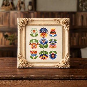 Puede incluir: Un diseño floral en punto de cruz enmarcado. La obra de arte presenta nueve flores estilizadas coloridas en tonos de azul, naranja, amarillo y verde. El marco es de color beige claro con detalles de rosas en las esquinas, sobre una superficie de madera oscura.