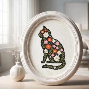 Puede incluir: Una obra de arte de punto de cruz enmarcada en óvalo blanco con un diseño de gato floral. La silueta del gato está llena de flores coloridas en tonos naranja, rosa, blanco y verde. La obra de arte se exhibe sobre un fondo claro.