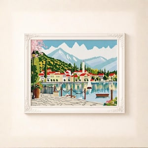 May include: A framed cross-stitch landscape showing a lakeside village with red-roofed buildings, a tall white tower, and snow-capped mountains. The artwork is in a white, ornate frame.