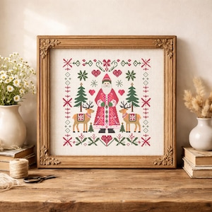 May include: A framed cross-stitch embroidery featuring a central figure of Santa Claus in a red coat, flanked by two reindeer and two evergreen trees. The design includes red hearts, green snowflakes, and geometric patterns on a white fabric background.